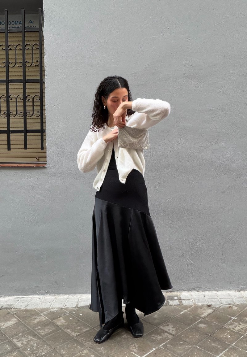 Witte gebreide cardigan over een zwarte mouwloze jurk met een uitlopende zoom. De outfit wordt aangevuld met zwarte schoenen en een zilveren, gestructureerde handtas.