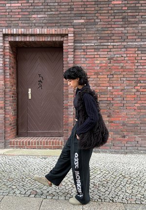 Persona con cabello negro que lleva un suéter negro, pantalones holgados con texto blanco y pantuflas, caminando por una calle de adoquines cerca de una pared de ladrillo y una puerta marrón.