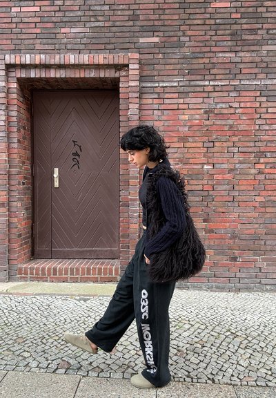 Persona con cabello negro que lleva un suéter negro, pantalones holgados con texto blanco y pantuflas, caminando por una calle de adoquines cerca de una pared de ladrillo y una puerta marrón.