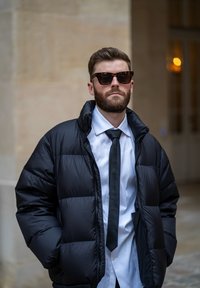 Fekete párnázott dzseki magas gallérral, fehér ing és fekete nyakkendő felett viselve. Sötét keretes napszemüveg egészíti ki a megjelenést.
