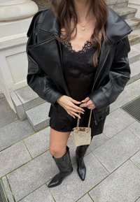 Giacca oversize in pelle nera sopra un vestito nero con bordi in pizzo, abbinata a stivali neri a punta sopra il ginocchio e una piccola borsa metallica.