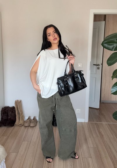 Mujer con cabello largo y negro lleva una blusa blanca sin mangas y pantalones sueltos color oliva, sosteniendo un bolso negro y gafas de sol, de pie en el interior.