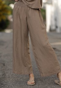 Pantaloni marroni leggeri e larghi con vita arricciata, gambe dritte e una texture liscia, abbinati a sandali semplici.