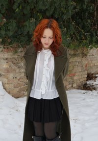 Jeune femme aux cheveux roux portant un manteau olive, une blouse blanche à volants, une jupe noire et des bottes, debout sur la neige près d'un mur de briques avec du lierre.