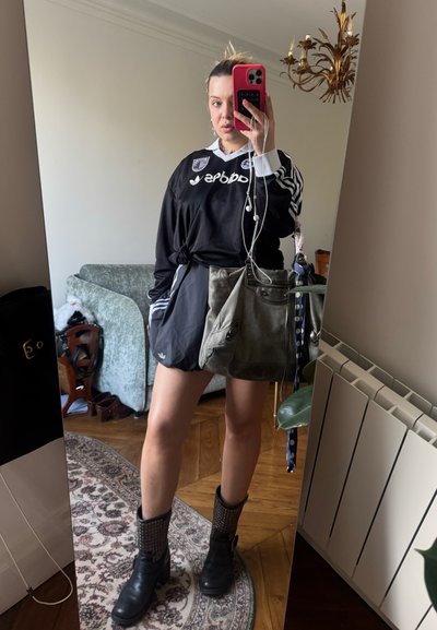 Jersey deportivo negro con rayas blancas, pantalones cortos negros, botas de tobillo negras texturizadas y una bolsa gris. Fondo de espejo reflectante.