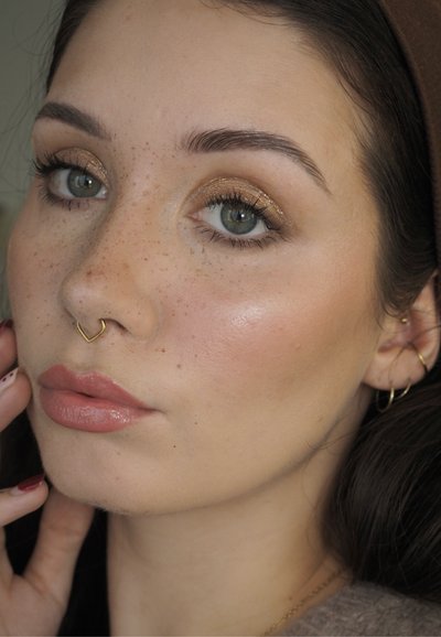 Primer plano de un rostro de piel clara con pecas, sombra de ojos dorada, piercing en el septum y labios nude brillantes. Se ven discretos pendientes.