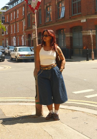 Culottes de denim de cintura alta, combinados con una camiseta blanca. Zapatos marrones, tatuajes visibles. De pie al lado de un cartel y un poste.