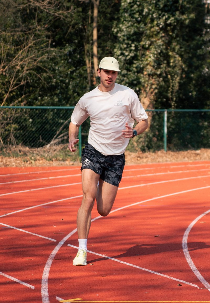 Man in shorts, t-shirt, and cap running on an outdoor red track with trees and fence in the background.