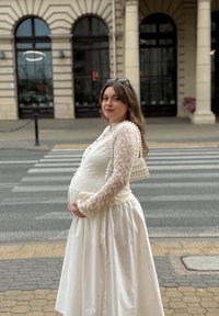 Noseča ženska v beli čipkasti obleki in s perlastim torbico stoji blizu prehoda za pešce v urbanem okolju s klasično zgradbo v ozadju.