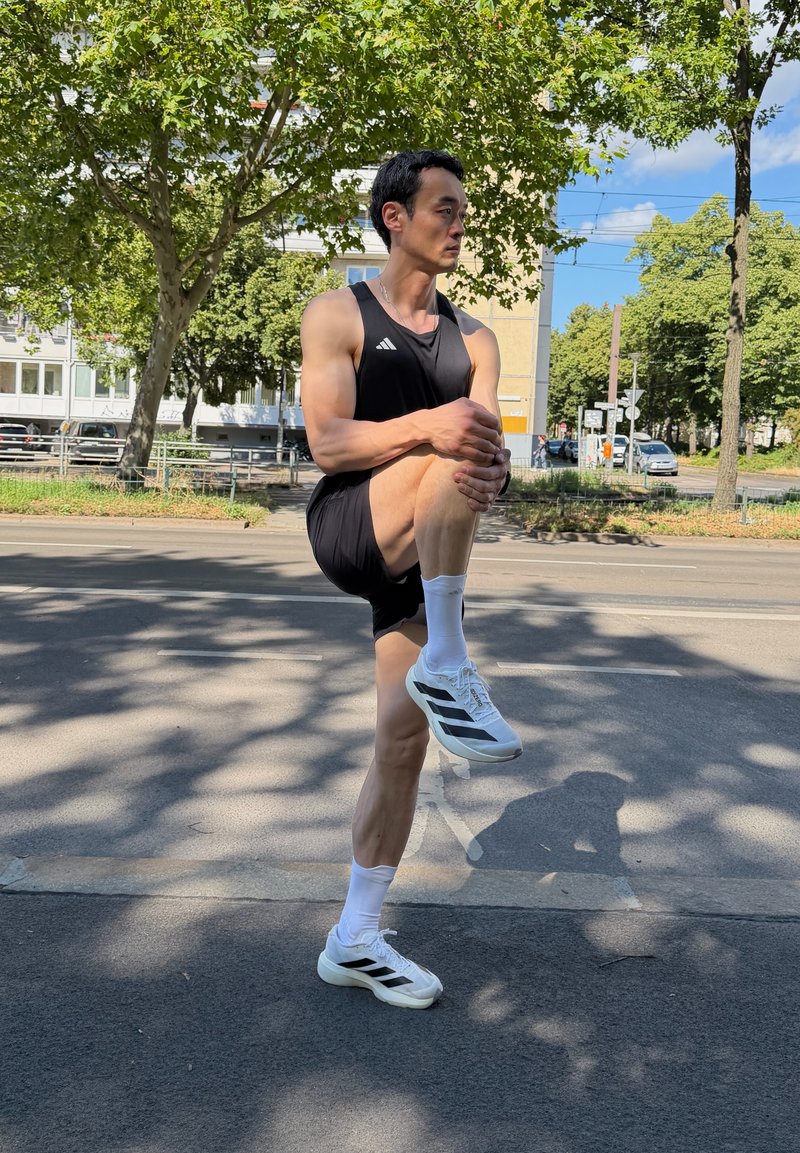 Man i svart sportkläder som stretchar ett ben på en stadsgata under träd en solig dag.