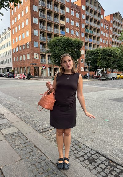 Vestido negro sin mangas con un diseño ajustado, zapatos negros texturizados y un bolso de mano tejido en naranja. Entorno urbano con edificios de ladrillo al fondo.