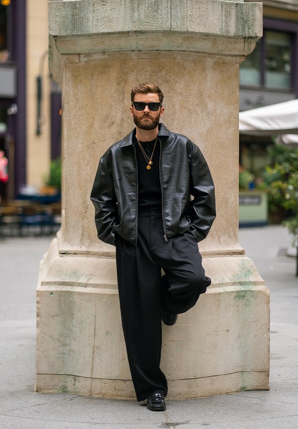 Un homme barbu, portant des lunettes de soleil, une veste en cuir noire et un pantalon noir ample, s’appuie contre un pilier de pierre dans une rue de la ville.
