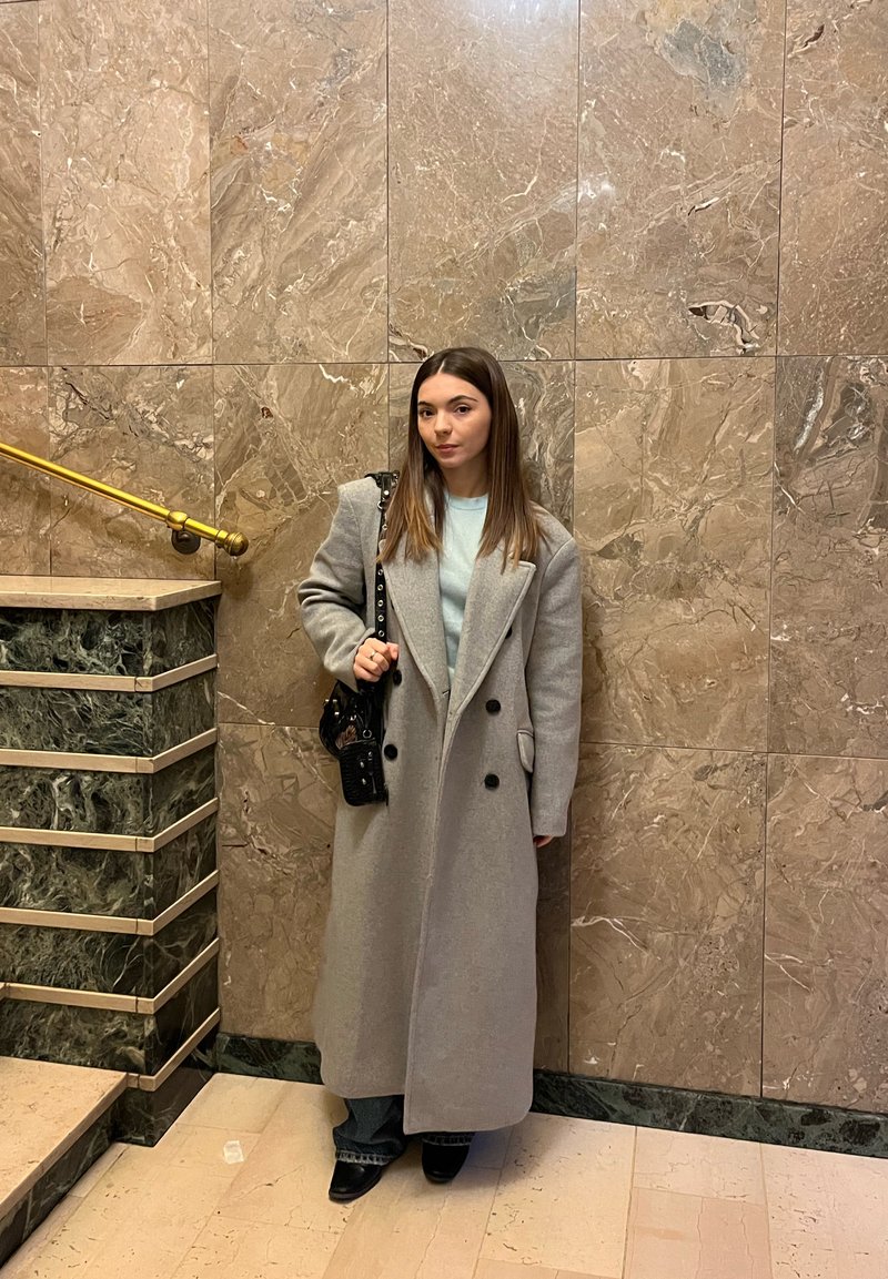 Palton lung gri din lână cu nasturi negri dubli și reveruri supradimensionate, asortat cu un top bleu deschis și blugi închiși la culoare. Geantă neagră.