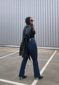 Manto trench de couro preto, top cropped preto ajustado, jeans flare azul de cintura alta e saltos altos pretos contra um fundo cinza corrugado.