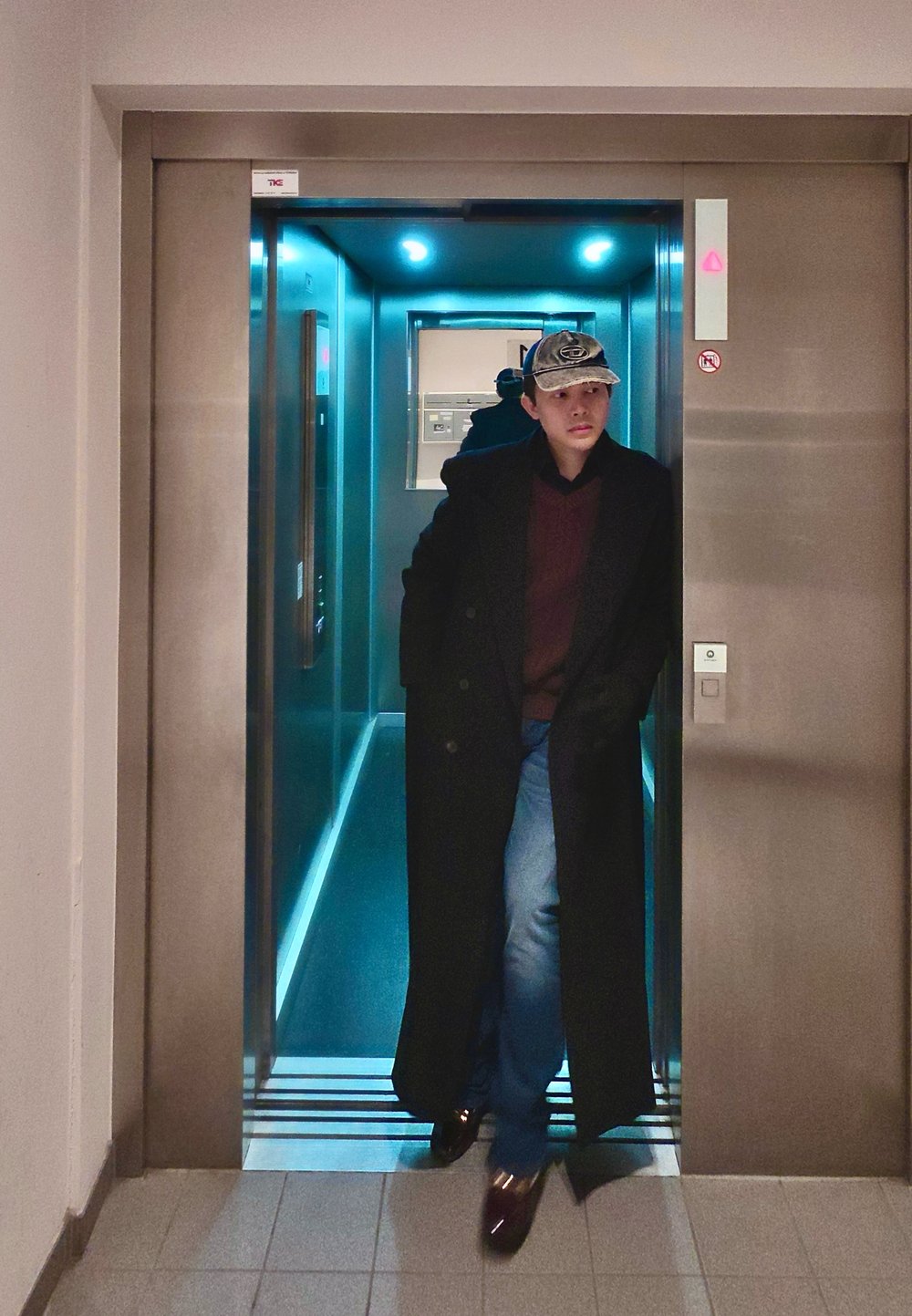 Man die een lange zwarte jas, een rode trui, jeans en een pet draagt en een verlichte lift met reflecterende muren uitloopt.