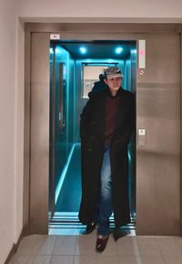 Man die een lange zwarte jas, een rode trui, jeans en een pet draagt en een verlichte lift met reflecterende muren uitloopt.
