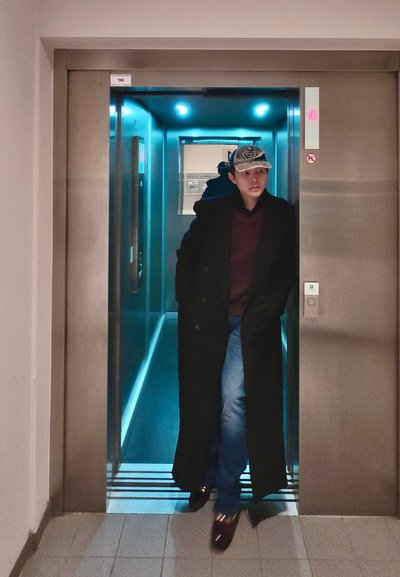 Hombre con un abrigo largo negro, suéter rojo, vaqueros y una gorra saliendo de un ascensor iluminado con paredes reflectantes.