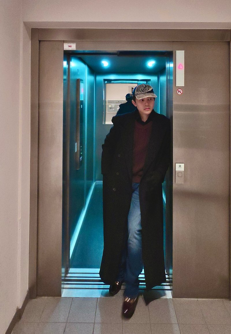Mand iført en lang sort frakke, rød sweater, jeans og en kasket, der går ud af en oplyst elevator med reflekterende vægge.