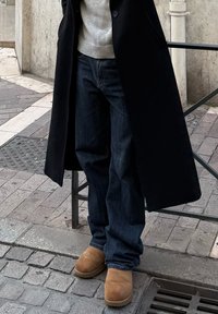 Personne portant un jean foncé, des bottes en daim marron clair, un pull gris et un long manteau noir, debout sur une rue pavée en pierre près d'une rambarde en métal.