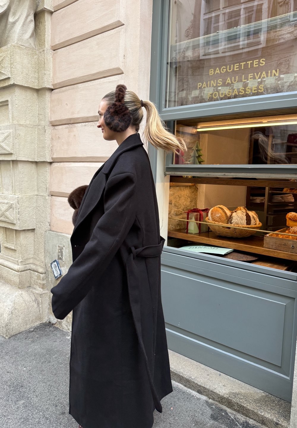 Long manteau noir avec de grands revers, taille cintrée et grandes poches. Cache-oreilles en peluche marron, coiffure lisse, et arrière-plan d'une vitrine de boulangerie.