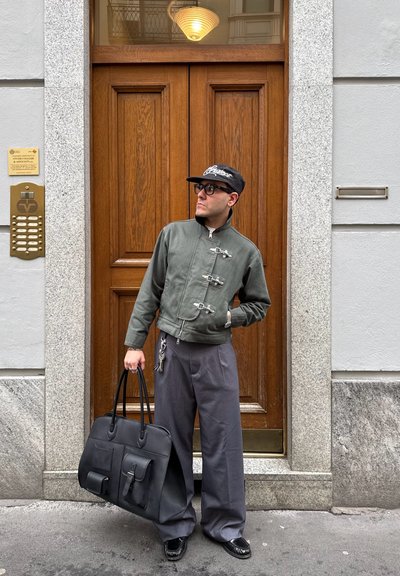 Hombre que lleva una chaqueta verde y una gorra negra sosteniendo una gran bolsa negra, de pie frente a una puerta de madera con marco de granito en una calle de la ciudad.