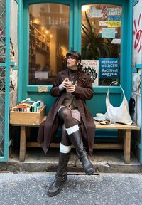 Brun frakk, rutete skjørt og brune høye støvler med hvite detaljer. Sitter med en drink ved siden av en trekasse med bøker.