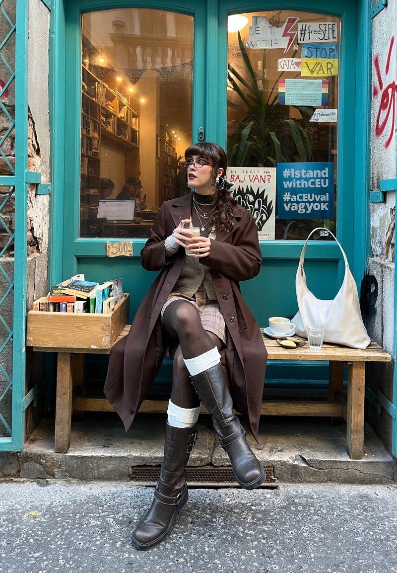 Brūna mēteļa, raibās svārku un brūnu zābaku līdz ceļiem ar baltām akcentiem. Sēžot ar dzērienu blakus koka kastei ar grāmatām.