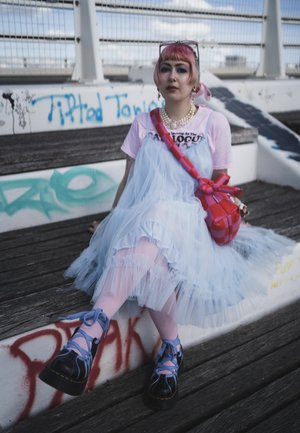 Persona con cabello rosa y maquillaje, vistiendo un vestido de tul azul claro, camisa rosa, zapatos de plataforma y un bolso rojo a cuadros sentada en unos escalones de madera cubiertos de grafitis.