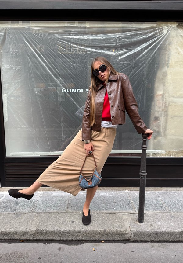 Une femme portant des lunettes de soleil, une veste en cuir marron et une jupe beige pose sur un trottoir en ville, tenant un sac à main bleu à côté d’un poteau métallique.