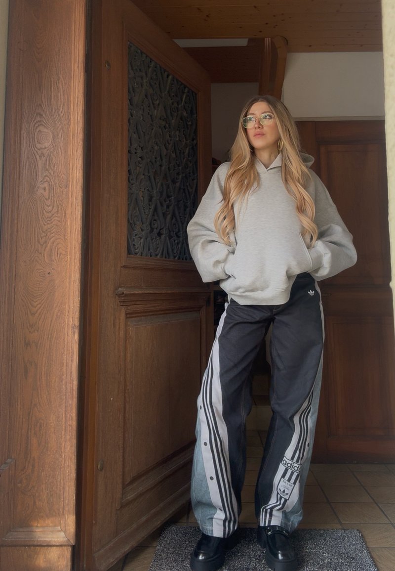 Young woman with long hair and glasses wearing gray hoodie and black Adidas track pants, standing indoors near a wooden door.