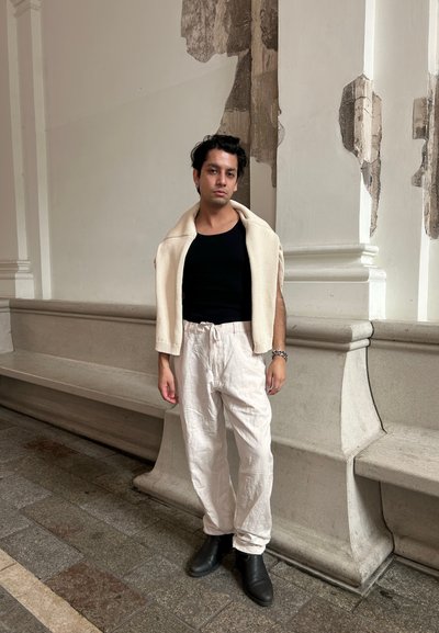 Suéter blanco de punto texturizado encima de una camiseta sin mangas negra, combinado con pantalones blancos holgados y botas de tobillo negras, con un fondo de piedra.