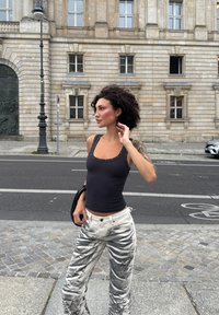 Top in cotone nero a costine con borchie decorative, abbinato a pantaloni bianchi con motivo astratto nero. La modella si trova all'aperto vicino a un edificio.