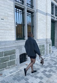Abrigo de lana negro grueso con cuello alto, combinado con una falda negra y medias transparentes. Bolso de hombro marrón, caminando sobre un pavimento de piedra.