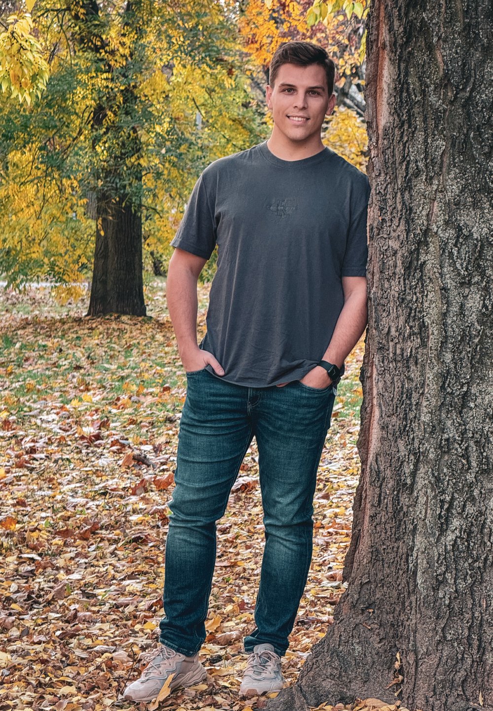 Lässiges graues T-Shirt, dunkelblaue Jeans und beige Sneaker. Steht gegen einen Baum mit strukturiertem Rinde, umgeben von herbstlichen Blättern.