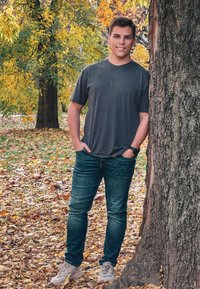 Lässiges graues T-Shirt, dunkelblaue Jeans und beige Sneaker. Steht gegen einen Baum mit strukturiertem Rinde, umgeben von herbstlichen Blättern.