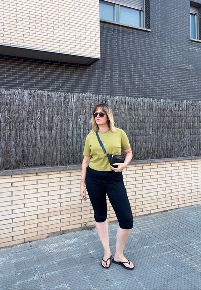 Mujer con cabello rubio que lleva gafas de sol, camiseta verde de manga corta, pantalones capri negros y sandalias negras, de pie en una acera de la ciudad.