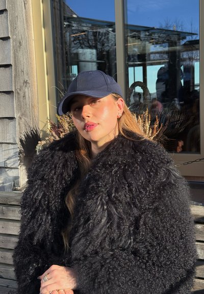 Joven mujer con gorra azul marino y abrigo negro rizado y grande sentada en un banco de madera al aire libre bajo la luz brillante del sol.