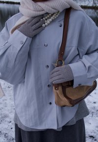 Personne portant des gants gris, un manteau bleu clair, une écharpe crème, un collier de perles, et tenant un sac à main marron texturé dans un décor extérieur enneigé.