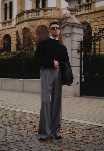 Suéter negro, pantalones anchos gris y gafas de sol oscuras; sosteniendo una bolsa negra. Pavimento texturizado con hojas caídas en el fondo.