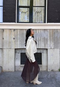 Giacca di pelle bianca con frange, abito maxi trasparente marrone, stivaletti beige. In piedi contro un muro grigio testurizzato; due finestre verdi sopra.