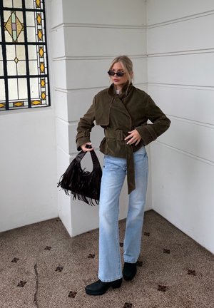 Mujer con chaqueta marrón de gran tamaño con cinturón, vaqueros azul claro, botas negras y gafas de sol, sosteniendo un bolso negro con flecos junto a una ventana de vitrales.