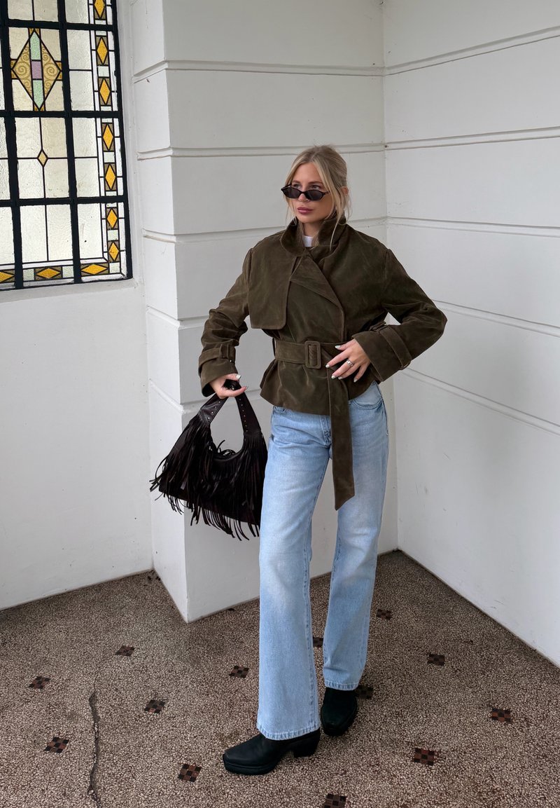 Femme portant une veste marron surdimensionnée ceinturée, un jean bleu clair, des bottes noires et des lunettes de soleil, tenant un sac à main noir à franges près d'une fenêtre en vitrail.