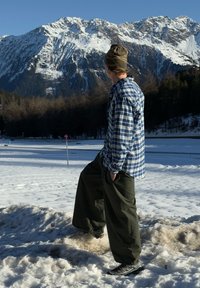 Chemise en flanelle à carreaux bleu et blanc, pantalon ample vert olive, et chaussures noires ; toile de fond de montagnes enneigées sous un ciel bleu clair.