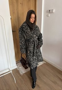 Manteau imprimé léopard avec une ceinture à la taille, en matière douce ; accompagné d'une capuche en tricot marron et de gants en cuir noir, tenant un sac bordeaux.