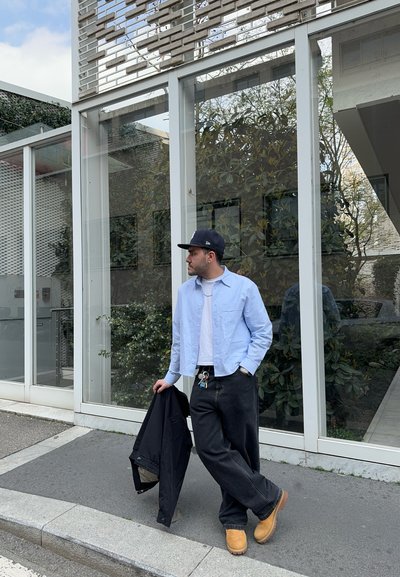 Camisa de botones celeste sobre una camiseta blanca, jeans holgados negros, zuecos marrones, sosteniendo una chaqueta negra, con vegetación y arquitectura moderna de fondo.