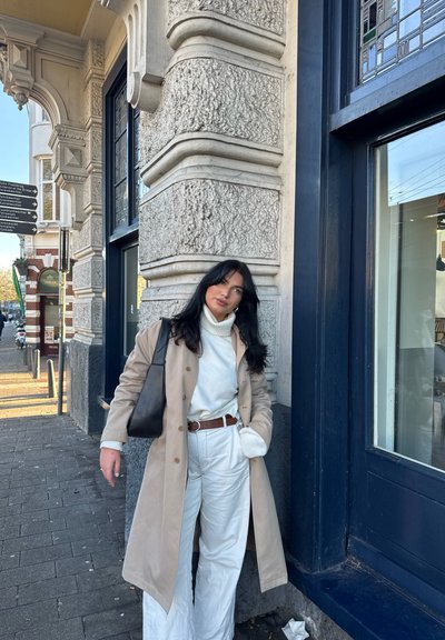 Trench beige sobre un cuello de tortuga blanco y pantalones anchos blancos, complementado con un cinturón marrón y un bolso negro. Fondo de un edificio característico.