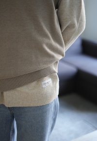 Beige sweater with a soft texture, ribbed hem, and a label at the back. Paired with light blue denim jeans.