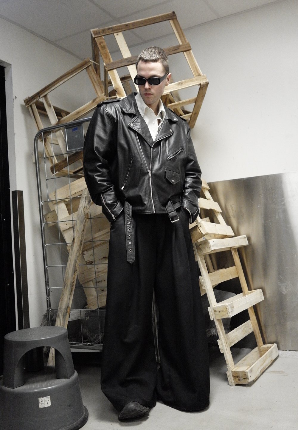 Veste de motard en cuir noir avec des fermetures éclair argentées, associée à un pantalon large noir avec un détail de sangle. Accessoires : lunettes de soleil.