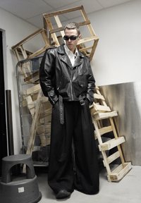 Veste de motard en cuir noir avec des fermetures éclair argentées, associée à un pantalon large noir avec un détail de sangle. Accessoires : lunettes de soleil.