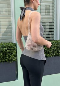 Sac à main translucide orné de perles avec une forme structurée, tenu par une personne portant un haut gris sans manches et un pantalon noir, se tenant devant un décor végétal.